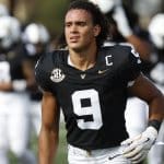 Football player in black jersey with number 9 and captain's C on his chest, on the field with teammates in the background.
